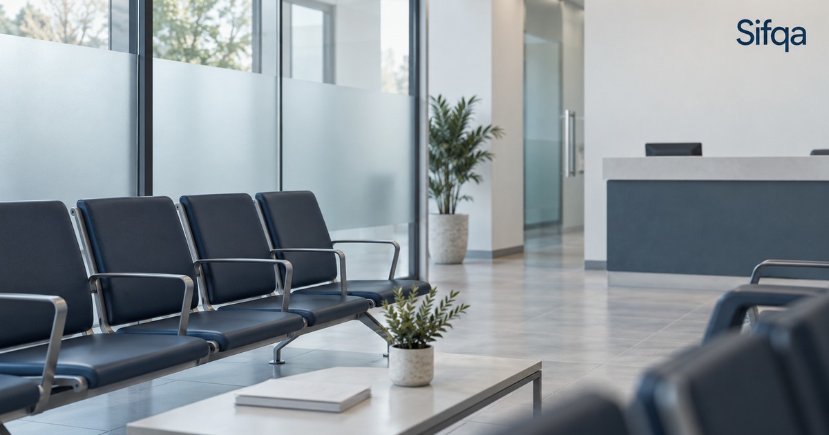 An empty waiting room with rows of dark blue chairs, a low table and plants beside a reception desk.