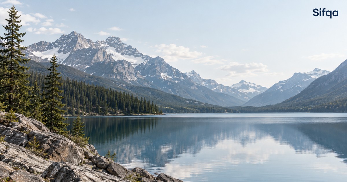 A calm lake reflecting snow-capped mountains and pine forest under a clear sky.