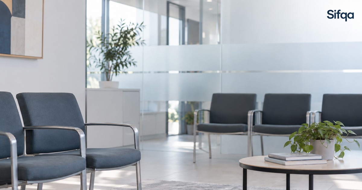 An empty waiting room with rows of grey chairs, a low table and plants beside frosted glass partitions.